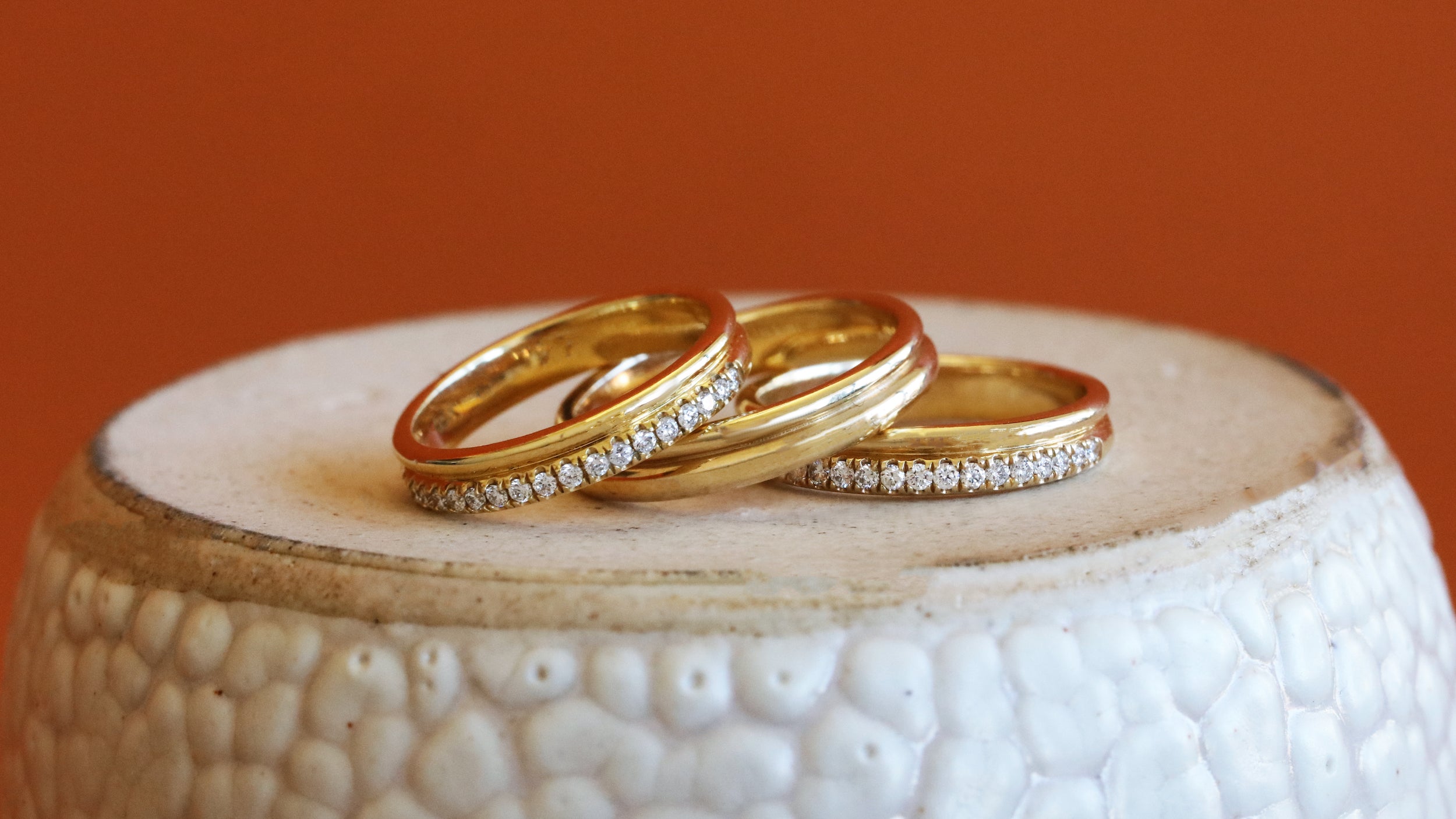 Lifestyle stacked front view of Duo Band / Pave Round Lab Diamonds, Duo Band, and Duo Band / Pave Round Natural Diamonds all in 14k yellow gold on a ceramic with orange background