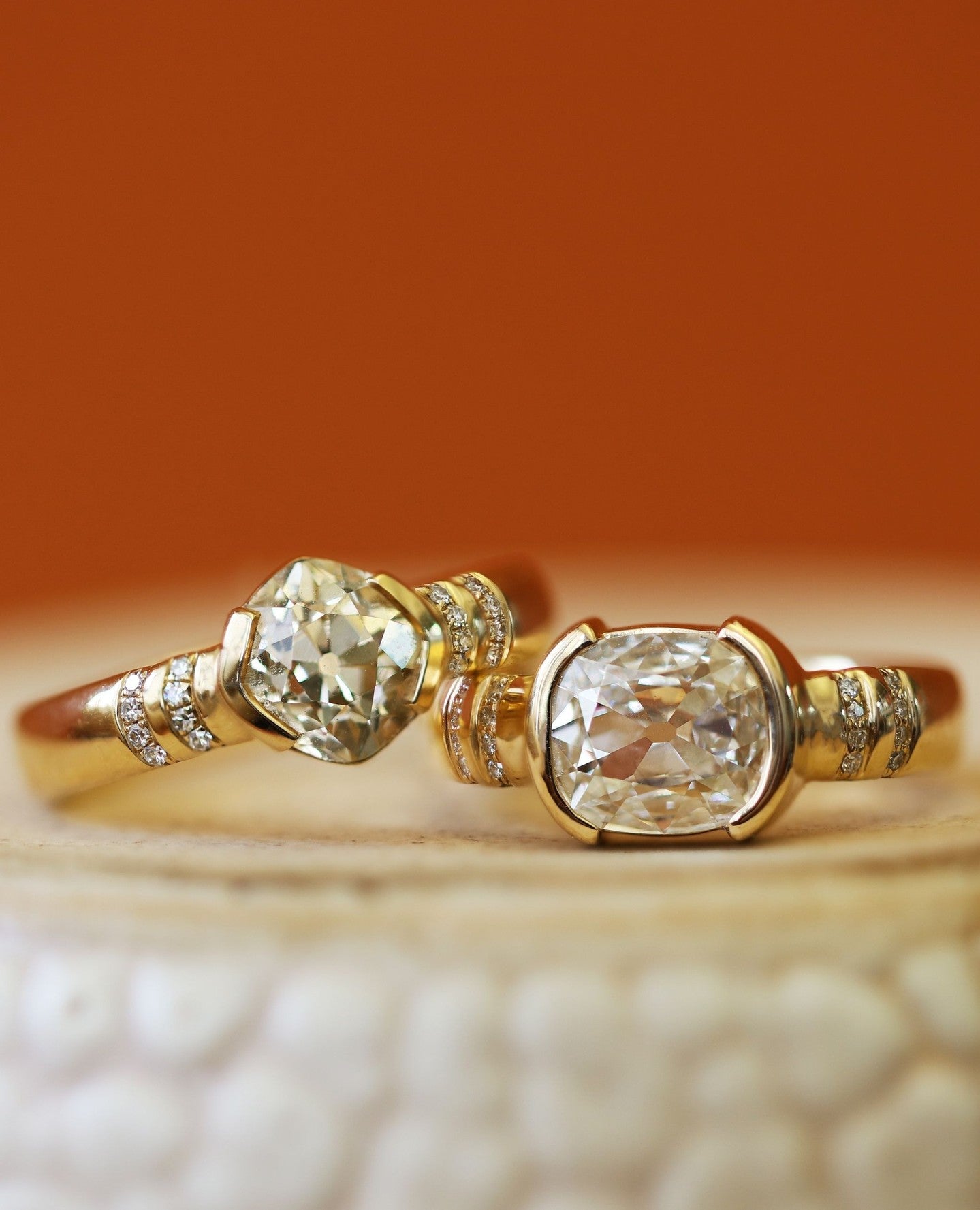 Stacked view of Dobbin Ring / Old Mine Natural Diamond 1.44ct and Dobbin Ring / Antique Oval Cushion Diamond 1.28ct by Goldpoint Jewelry on a ceramic with blurred orange background