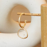 Lifestyle front view of Amorphous Pearl Bead Charm / Large Circle by Goldpoint Jewelry on an earring on a wooden display with a ceramic in the background
