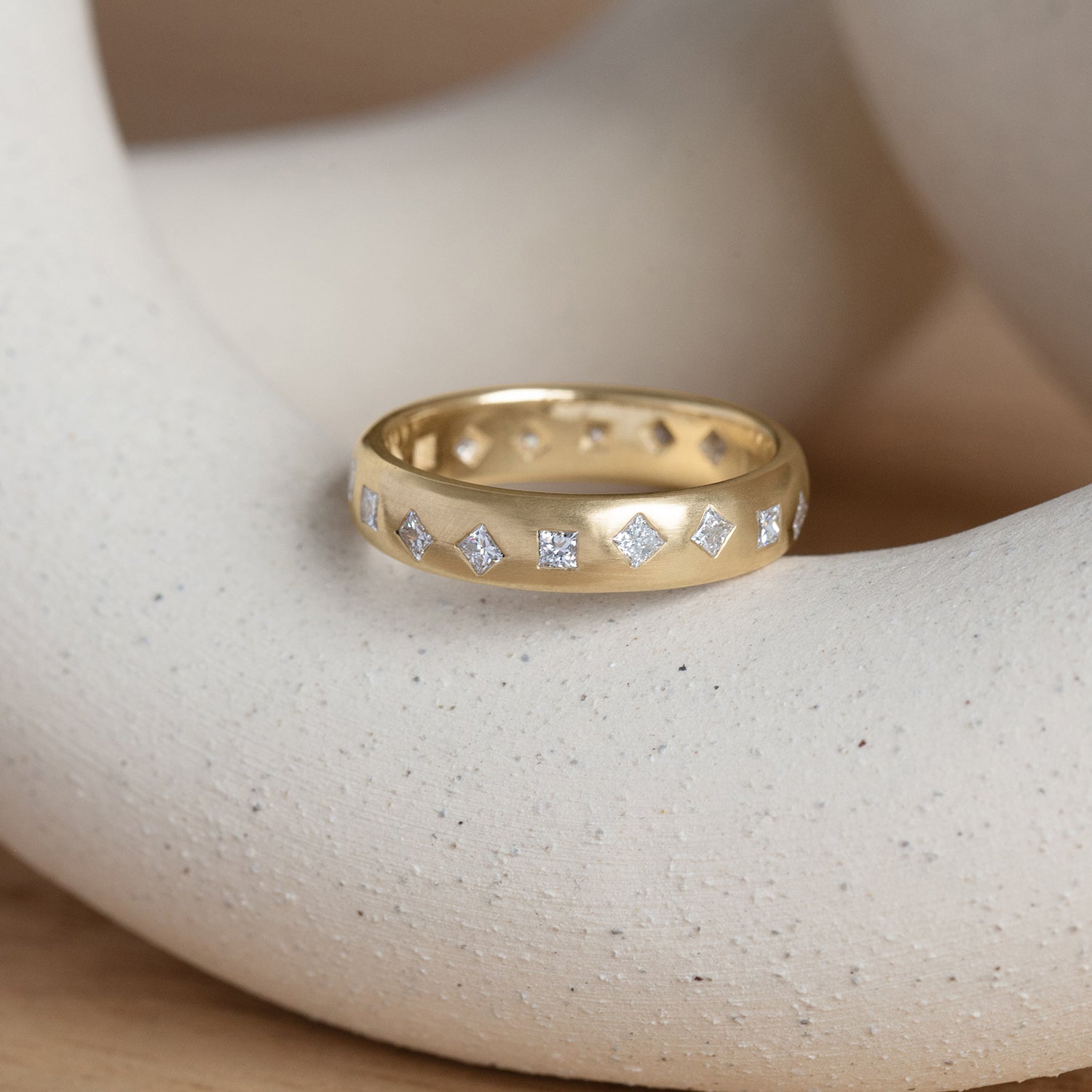 Lifestyle angled view of Dancing Band 5mm / Flush Princess Cut Lab Diamonds / in Yellow Gold on a ceramic with blurred natural backdrop