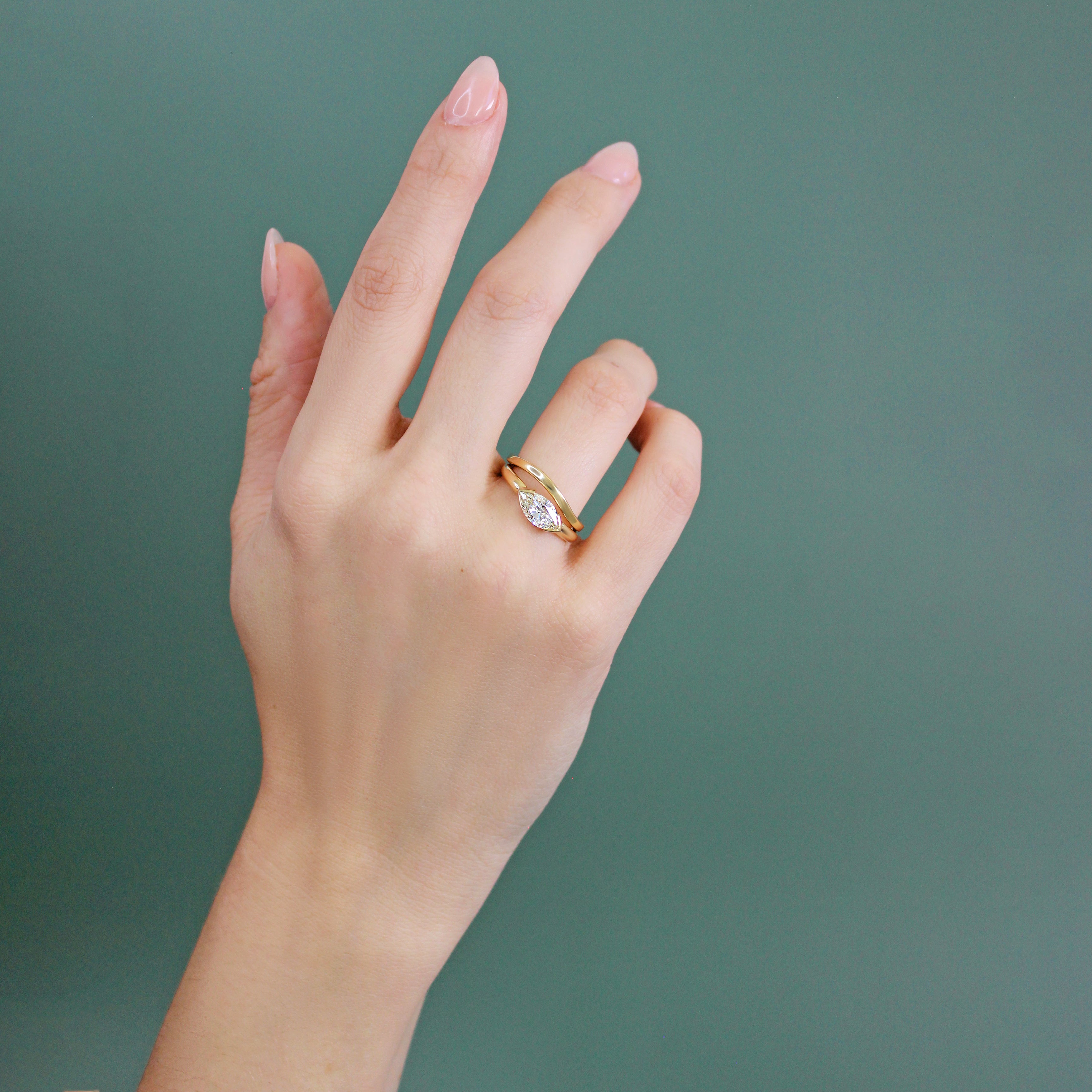 Model wears Flat Band Soft Curve / in 14k Yellow Gold stacked with an engagement by Goldpoint Jewelry on their ring finger with a teal background