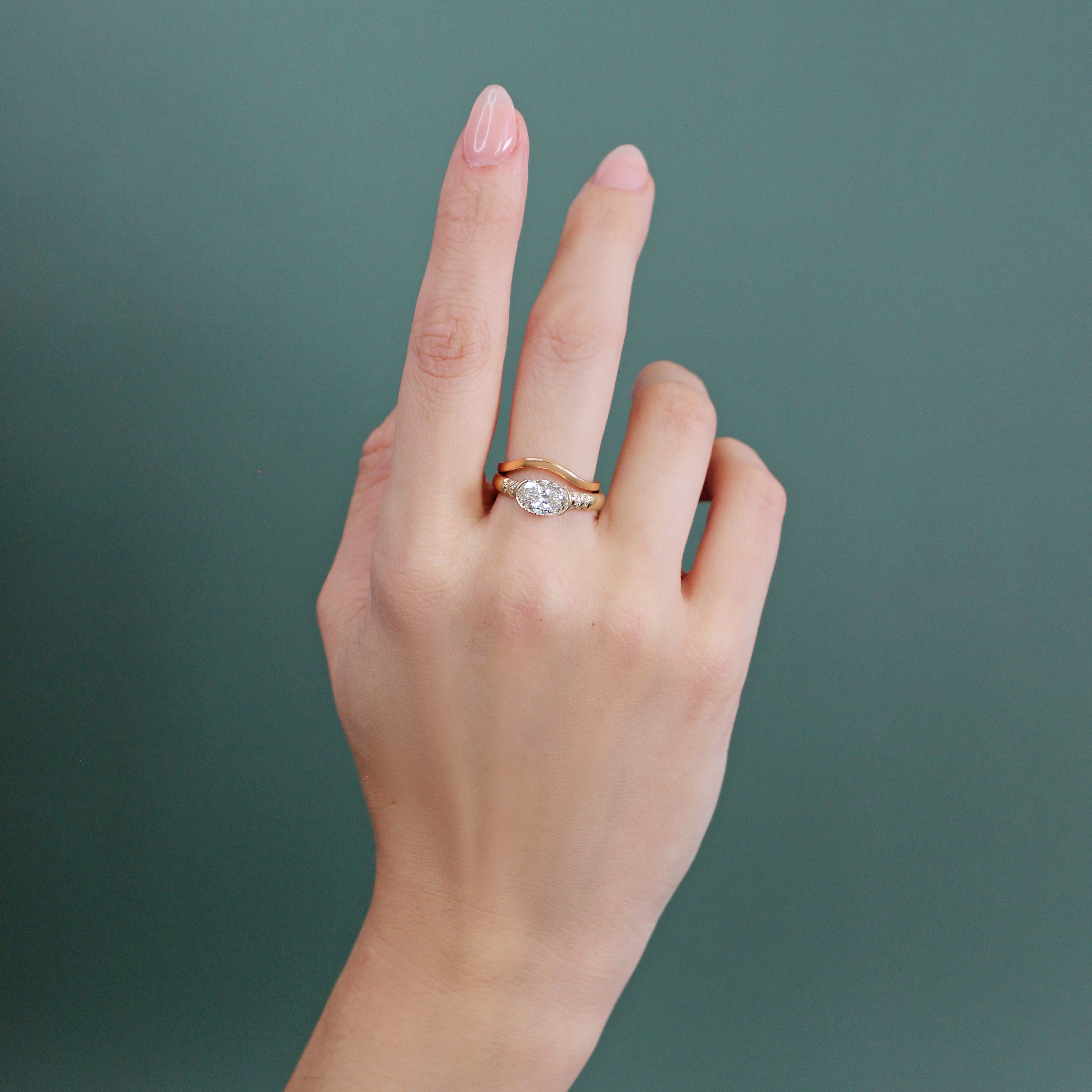 Model wears Flat Band Deep Curve in 14k yellow gold stacked with an engagement ring by Goldpoint Jewelry on their middle finger with a teal background