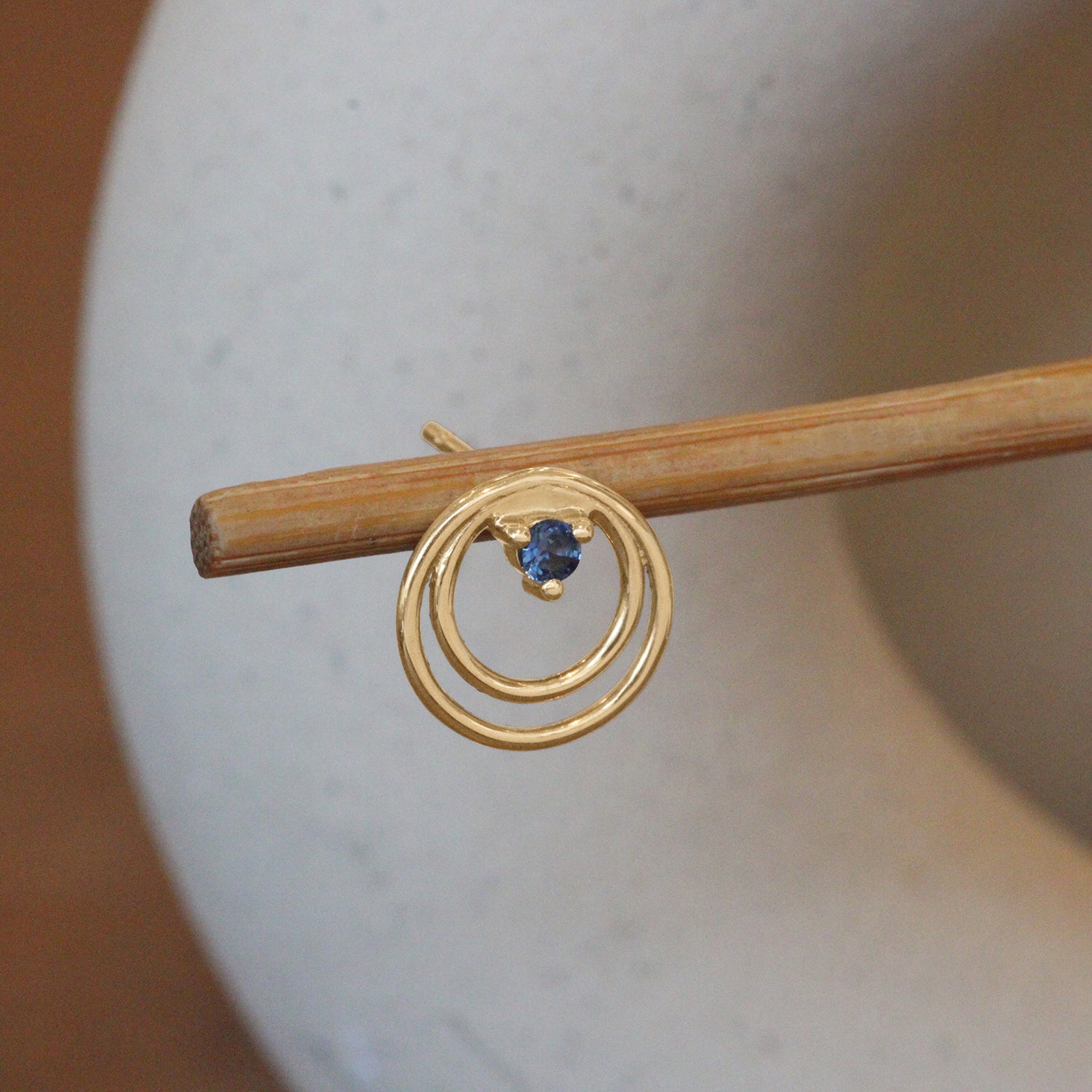 Lifestyle angled view of Ripple Earring / Round Sapphire by Goldpoint Jewelry on a wooden stand with a blurred background