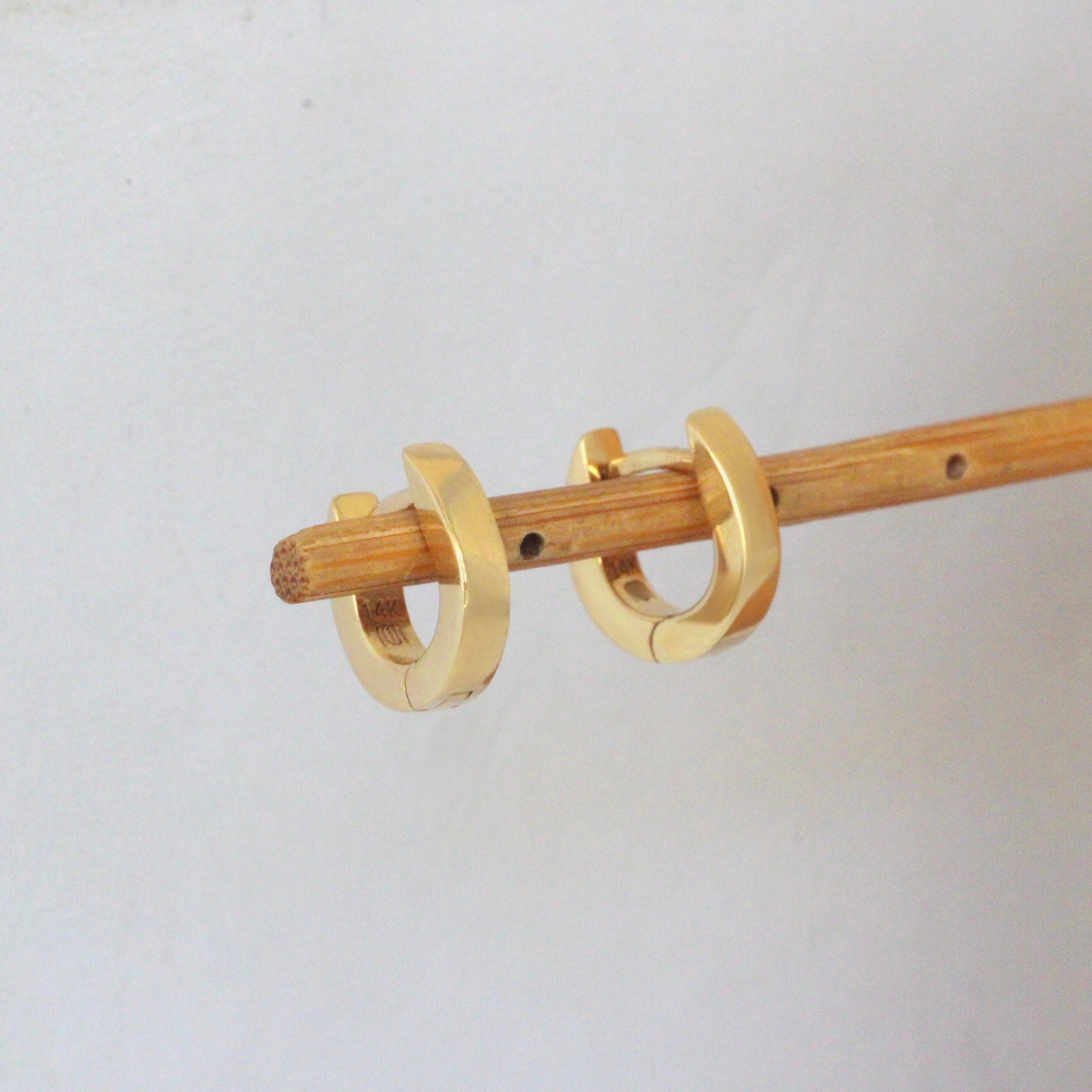 Lifestyle angled side view of Solid Huggie Hoops by Mon Pilar on a wooden stand with blurred backgroud