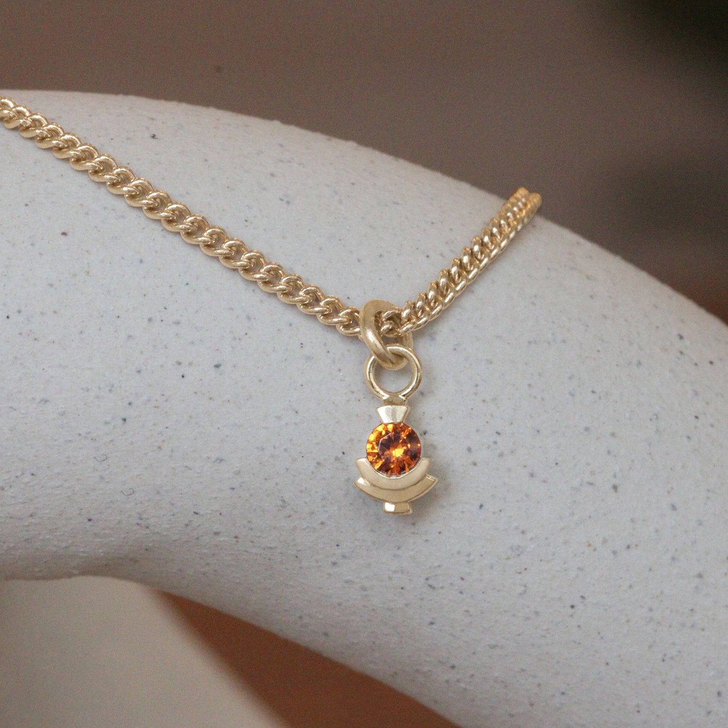 Lifestyle front view of Step Mazunte Charm / Orange Garnet on a Curb Chain by Goldpoint Jewelry on a ceramic with blurred background