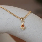 Lifestyle front view of Step Mazunte Charm / Orange Garnet on a Curb Chain by Goldpoint Jewelry on a ceramic with blurred background