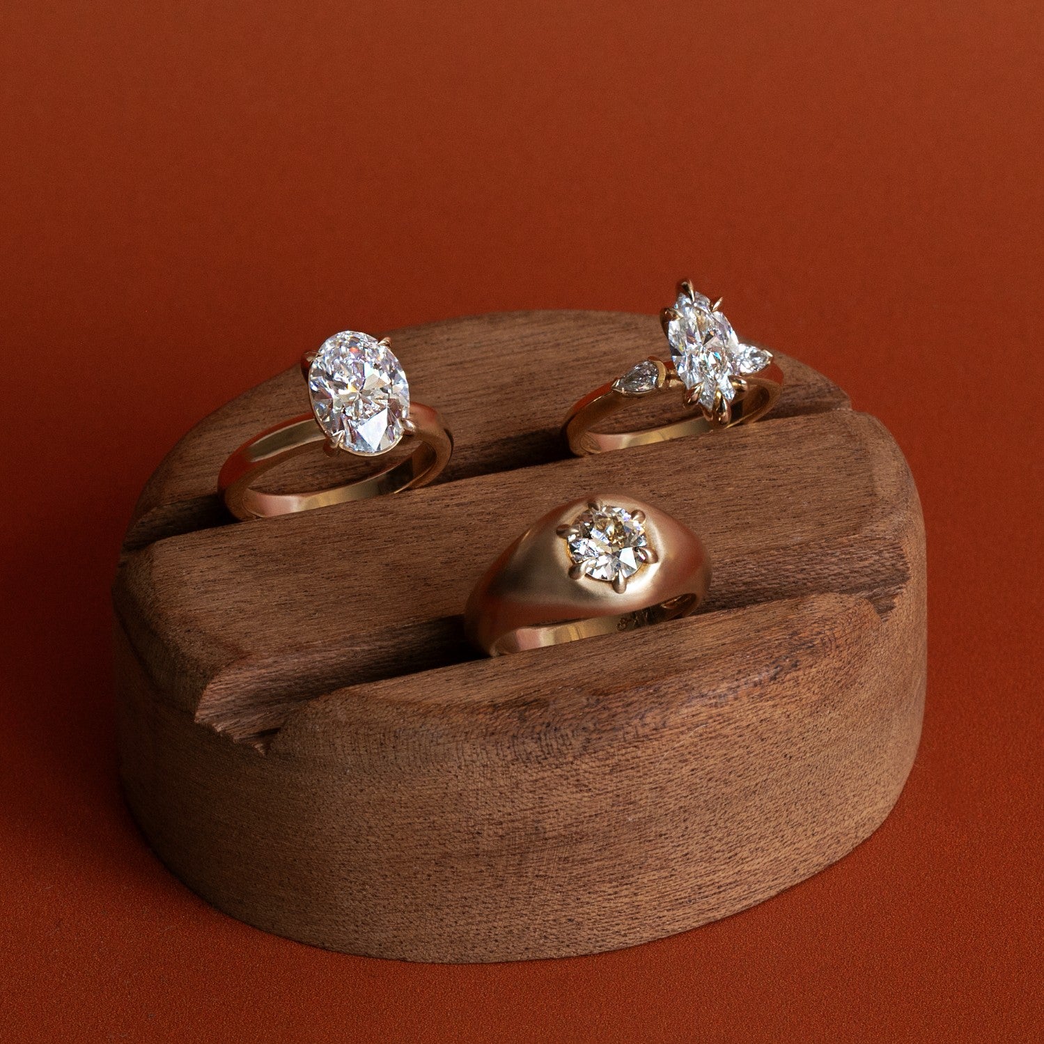 Three white diamond engagement rings by Goldpoint Jewelry on a wooden ring display with orange backdrop