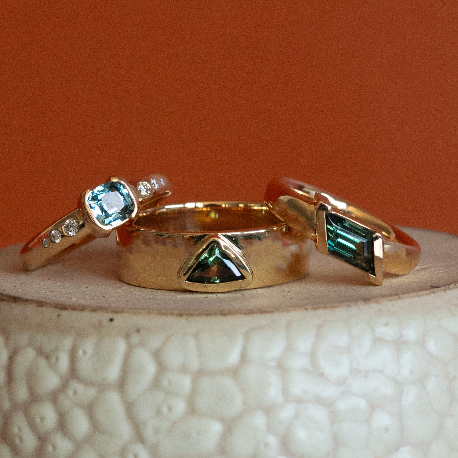 Three gemstone engagement rings by Goldpoint Jewelry stacked on eachother sitting on a ceramic with an orange backdrop