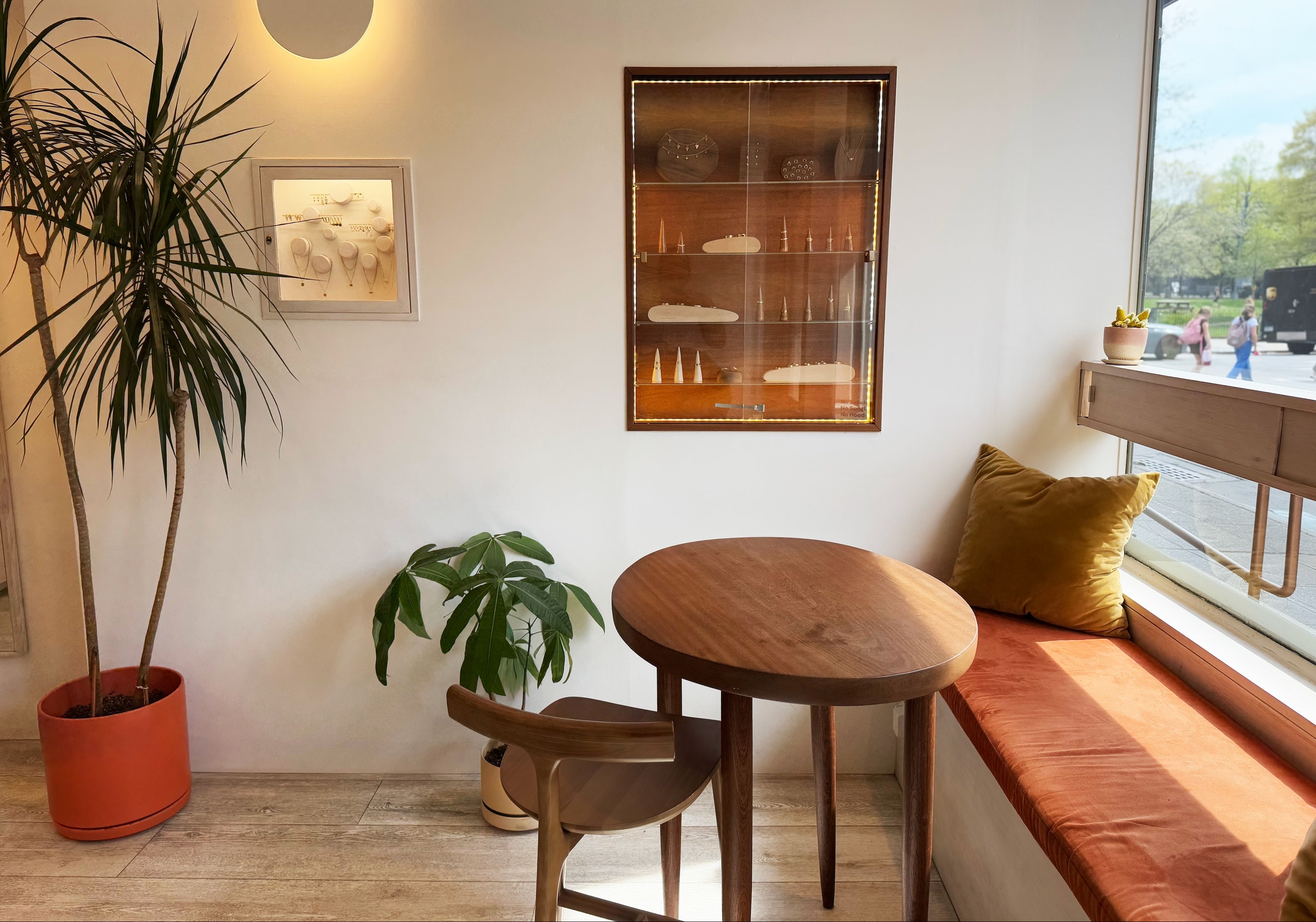Interiors of Goldpoint Jewelry in Greenpoint Brooklyn showcasing fine jewelry cases behind a small sitting area with a wooden table, chairs, and a bench by a window with plants.